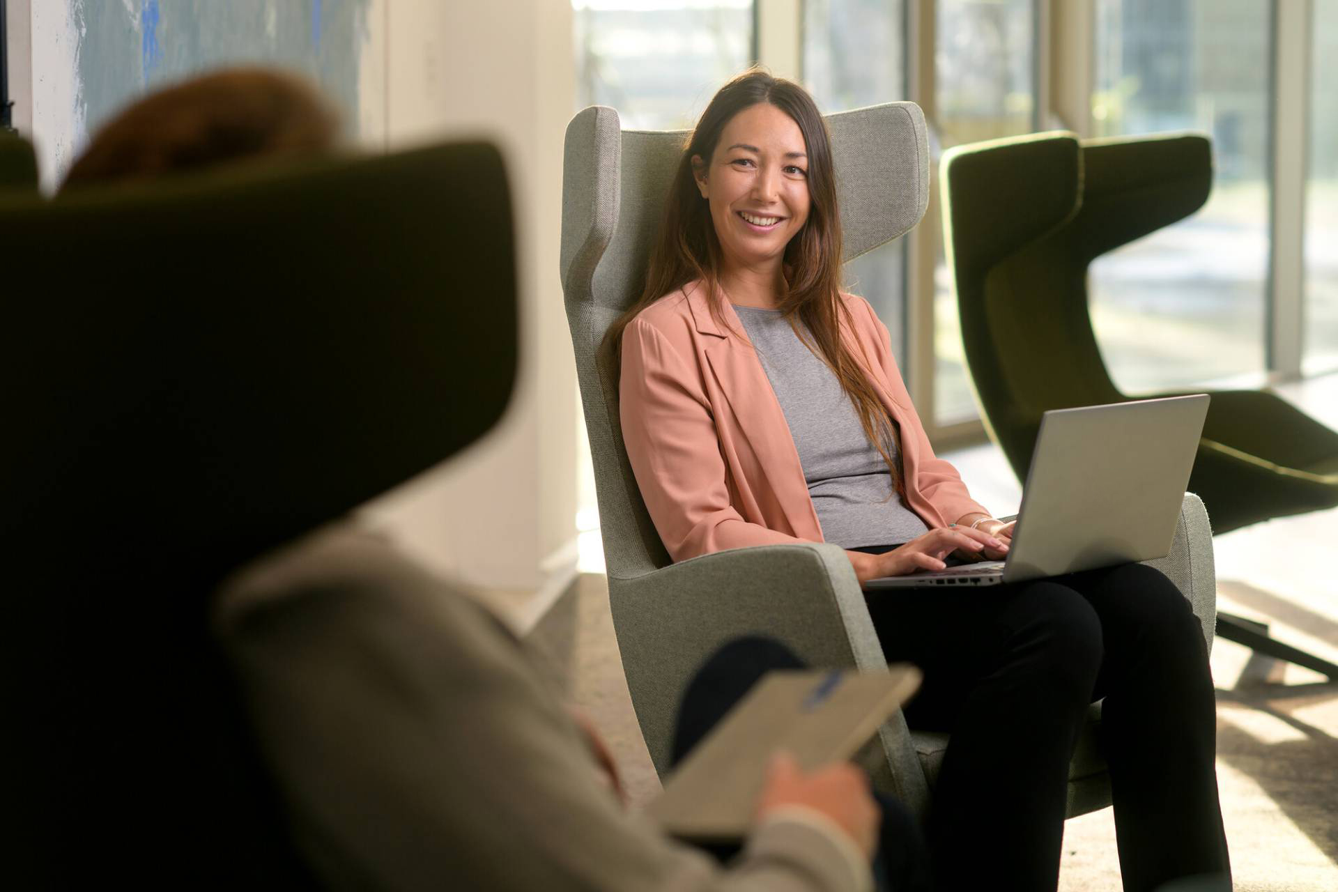 Medewerker van de Belastingdienst zit op een stoel achter haar laptop, lachend naar een collega