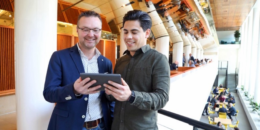 Dick Blaauw en een jonge frisse collega kijken lachend naar een tablet die ze samen vasthouden.