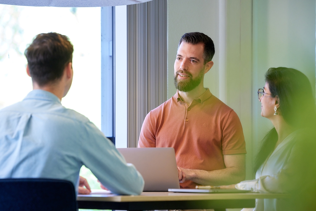 Twee mannelijke en een vrouwelijke collega staand aan tafel in gesprek