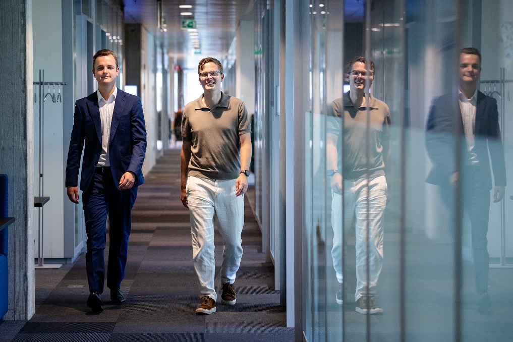 Twee mannen lopen door een kantoorhal en kijken richting de camera.