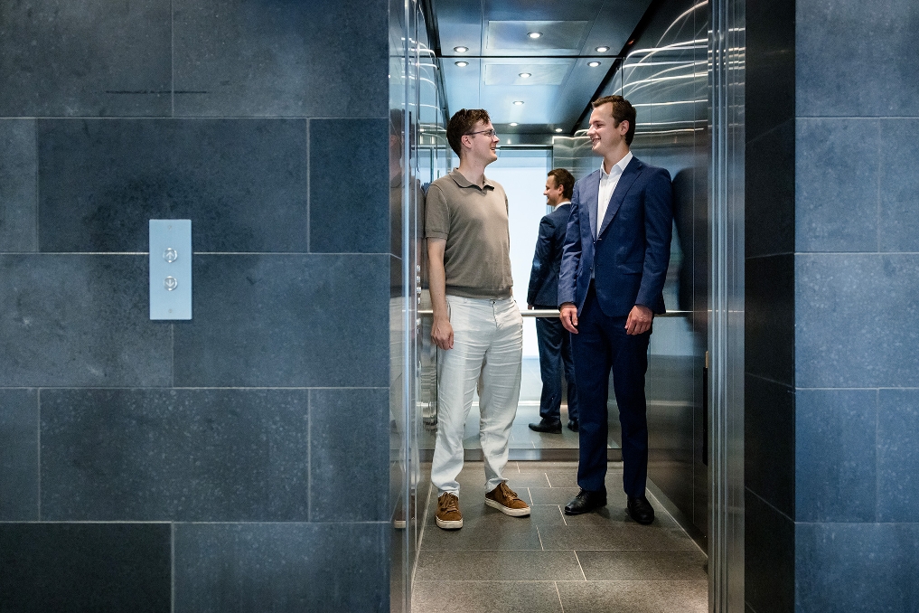 Twee mannen staan lachend in gesprek bij een lift in een kantoorgebouw.